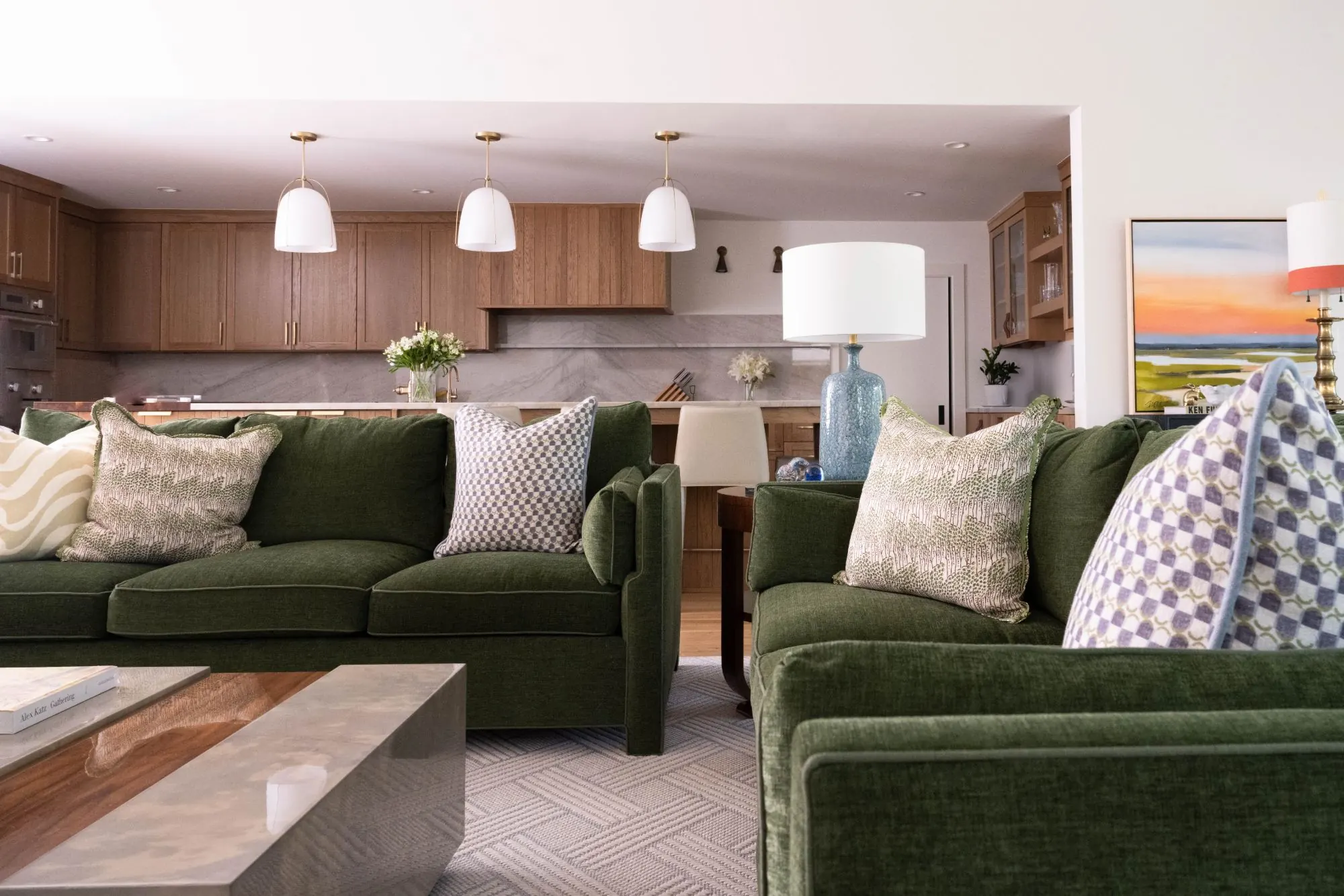 Living room with green velvet sofas, patterned pillows, and an open oak kitchen with brass pendants beyond