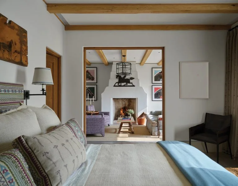 Bedroom with patterned pillows and a linen headboard, opening onto a sitting room with a white plaster fireplace and timber beams