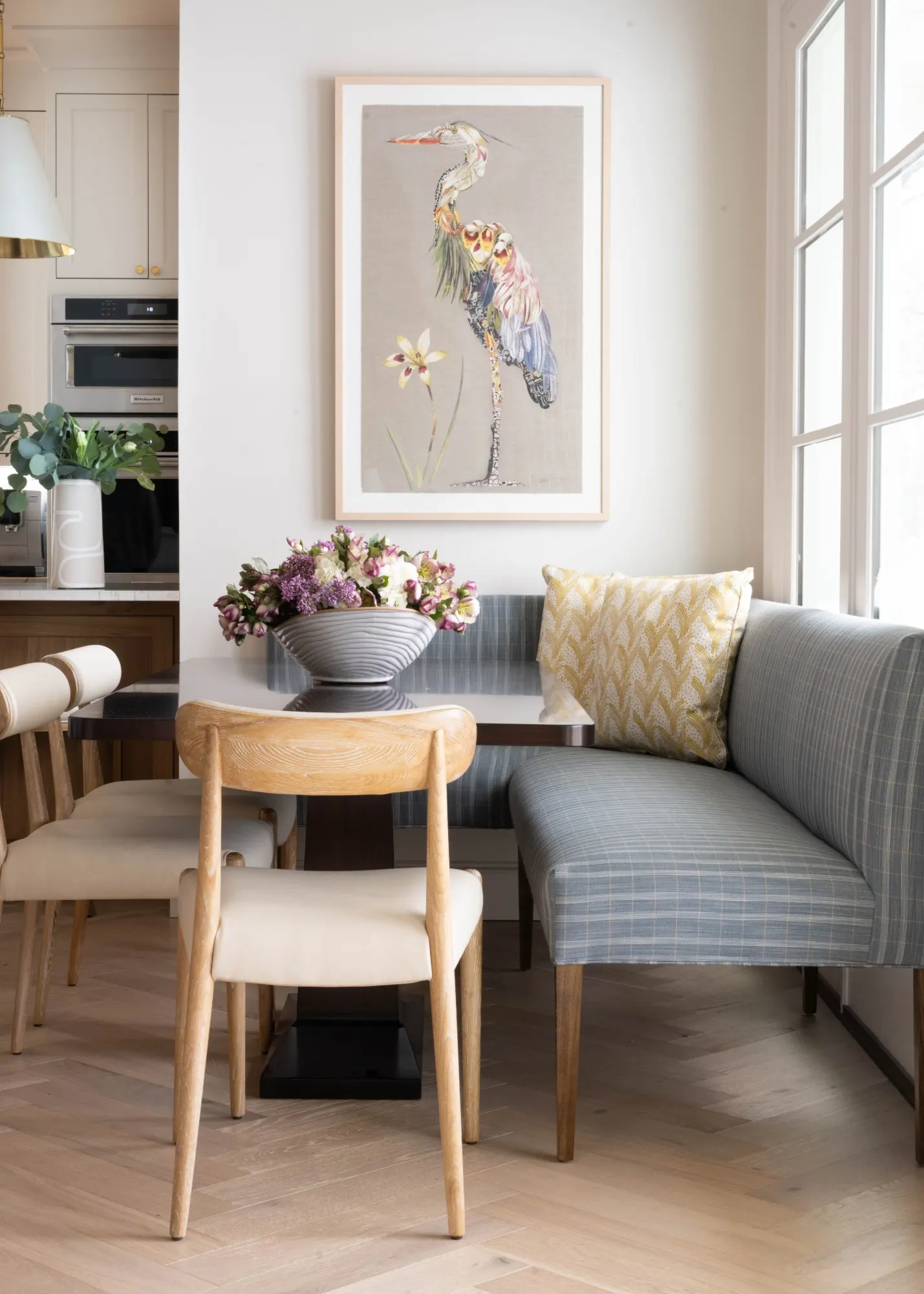 Breakfast nook with a blue plaid banquette, oak dining chairs, and a collage heron artwork