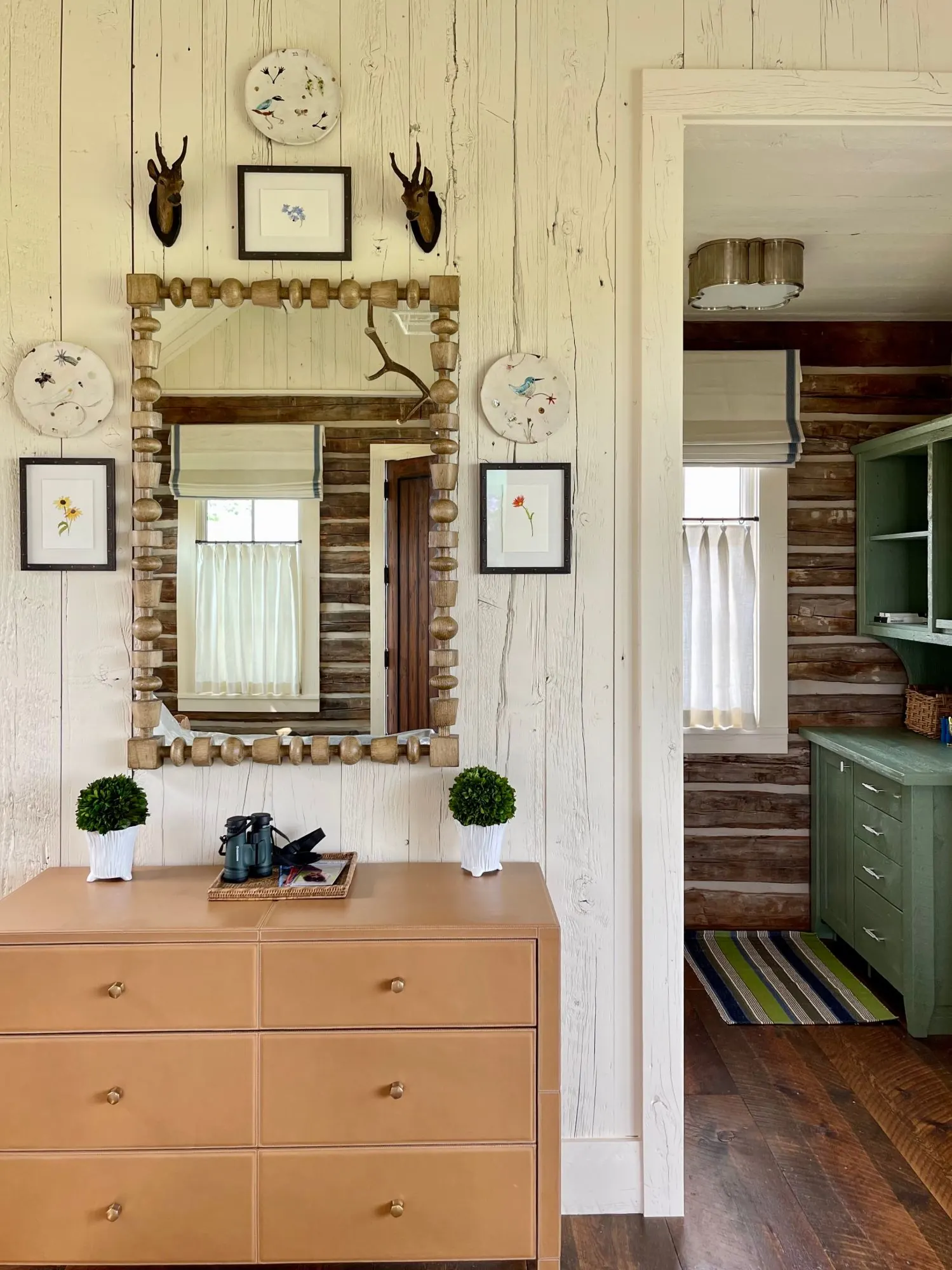 Whitewashed plank wall with a turned-wood mirror, botanical prints, and a leather chest, opening onto a log-walled room with green cabinetry