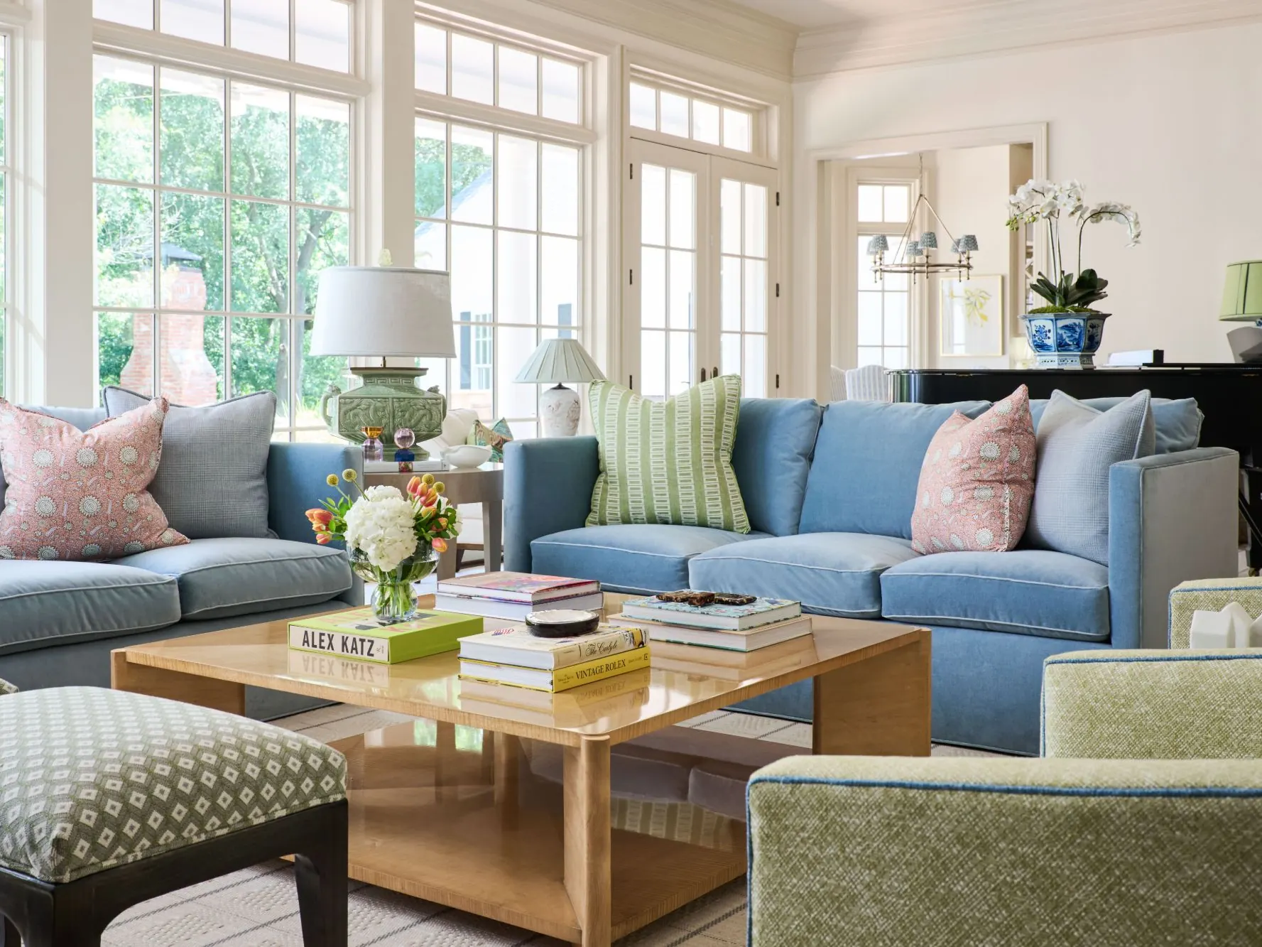 Sunlit living room with blue velvet sofas, pink and green pillows, and a maple coffee table before a wall of windows